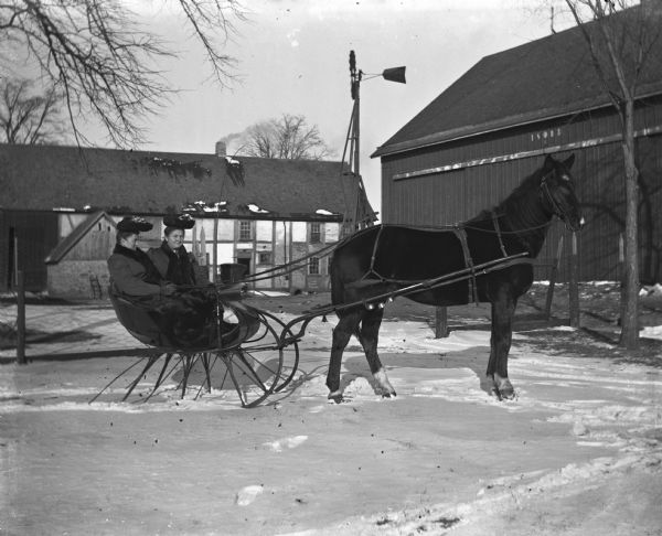 Ella and Ida Scholz sitting in a cutter (horse-drawn sleigh) in front of the Langholf half-timber home during winter.