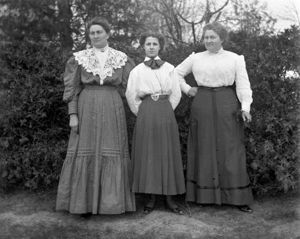 Outdoor group portrait of Ida Scholz, Edna Weigel, and Ella Scholz standing in front of a hedge.