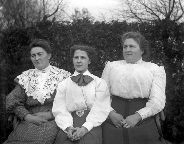 Outdoor portrait of Ida Scholz, Edna Weigel, and Ella Scholz sitting on a bench in front of a large hedge.