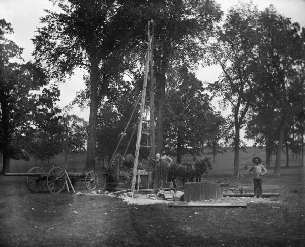 Two men drilling a well in the Will's woods using a cable tool drilling rig powered by a team of two horses. A wagon, various pipes, and other drilling equipment stand around the men.