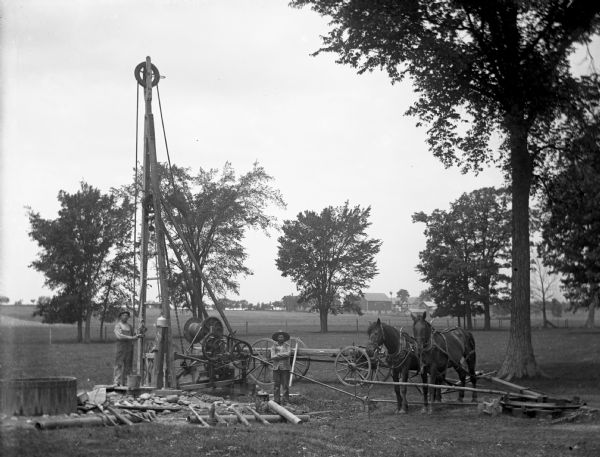 Two men drilling a well in the Will's woods using a cable tool drilling rig powered by a team of two horses. One man is standing next to the drill holding the bit, while the other man is standing nearby. A wagon, pipes, and various other drilling equipment are near the drill. A farmhouse and various farm buildings are in the far distance.