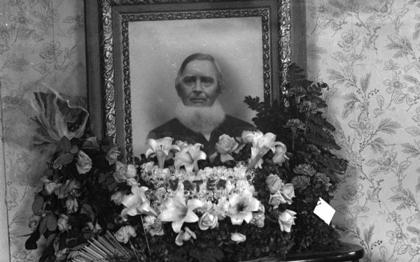 Floral funeral arrangement for William Jaeger. A large head and shoulders portrait of him is resting on two chairs behind floral arrangements, with "Vater" spelled out on top of the flowers in the center.