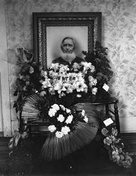 Funeral floral arrangement for William Jaeger. His framed portrait is sitting on two wooden chairs. He is wearing a suit and has a neckbeard. Flower arrangements are positioned on the chairs and on the floor in front of the portrait. The center floral arrangement has "Vater" spelled out on top of the flowers.