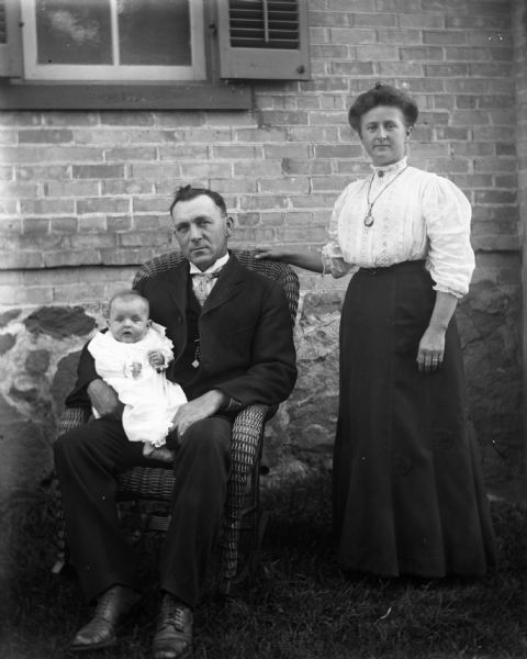 Outdoor group portrait of the John Lettho family posing in the yard with the brick house behind them. John is sitting in a wicker rocking chair holding his daughter, Viola, in his lap. Mrs. John Letto is standing beside her husband and is resting her hand on the back of the rocking chair.