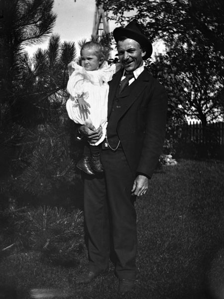 Outdoor portrait of Christ Bhend standing and holding his daughter, Ella, in a yard. In the background is the wooden support structure for what may be a windmill.