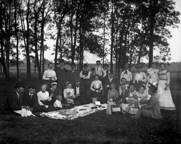 Group portrait of several neighbors and relatives picnicking together in the Grosnick's woods. The group sits around a large blanket that is laid out with various food items. Sarah Krueger stands in in the back row, first on the left.