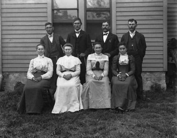 Outdoor family portrait of the Backhaus family posing in the yard of a house. Four men are standing in the back row while four women are sitting on chairs in the front row. Sarah Krueger is sitting second from the left.