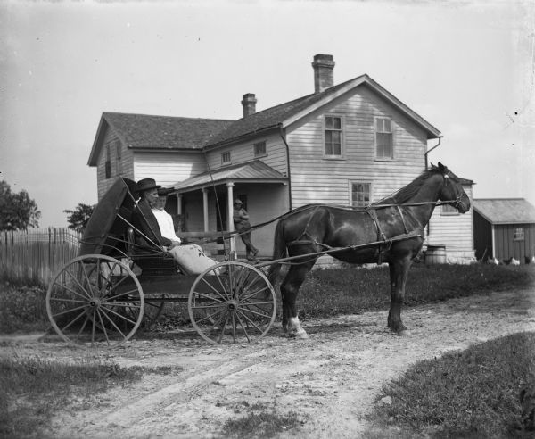 Albert Gennerman and his wife, Amanda, are sitting in a horse-drawn buggy in front of their home. On the front porch, an unidentified man is leaning against a support post. On the far right chickens are near a small outbuilding.