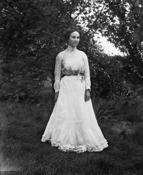 Outdoor portrait of Clara Krause (?) standing in the grass amongst several trees.