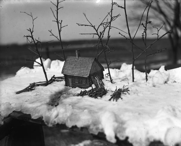 A small model of the first Krueger home, a log cabin located in Sugar Island. The model is sitting outdoors on a plank of wood covered in snow. Twigs are placed in the snow as trees and are used to make the fence and firewood piles.
