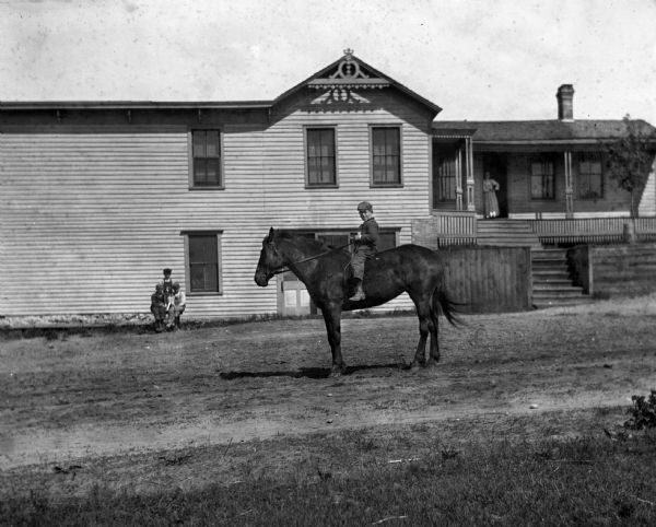 A young boy on horseback posing in front of the Hoffman home. Behind him posing near the side of the house is a group of boys posing with a dog. A woman is standing in the open doorway under the porch on the right.