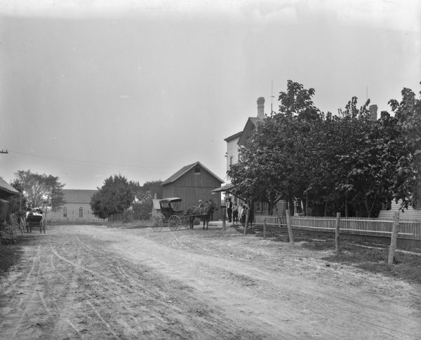 View down unpaved street towards the old part of town. Several horse-drawn carriages are parked on both sides of the street. A group of men, women and children are standing in front of a building on the right. On the left a group of people are sitting in a horse-drawn carriage.