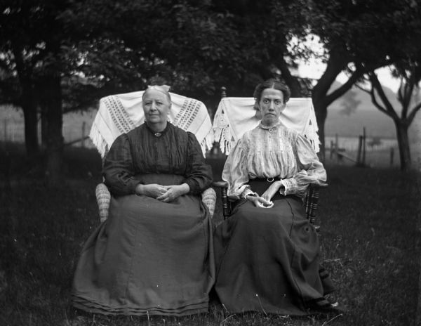 Outdoor portrait of two unidentified women posing in chairs with  shawls fastened to the backs. The two women are friends of Mr. and Mrs. August Krueger.
