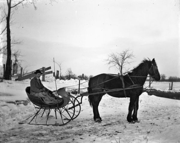 Winter scene with Joseph Langenbach sitting in a horse-drawn cutter (sleigh) at the end of the snowy driveway. In the background is a fence and a U.S. Mail box.