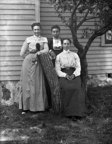 Outdoor portrait of three unidentified women posing at the side of a house next to a log stump that is fashioned into a chair. One of the women is sitting in the chair, while the other two women are standing behind her leaning on the back of the chair.