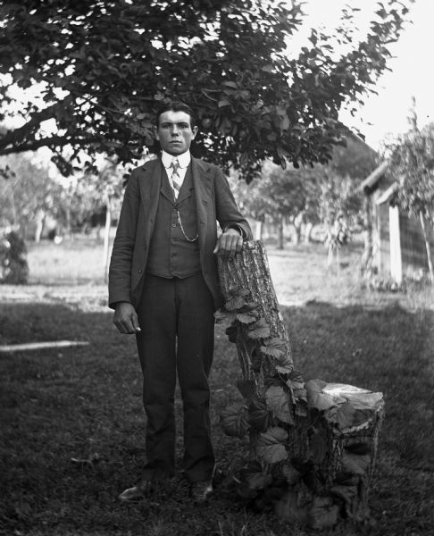 Outdoor portrait of Joseph Langenbach standing while resting his arm on the top of a log fashioned into a chair. A vine is growing on the stump. In the background are farm buildings.