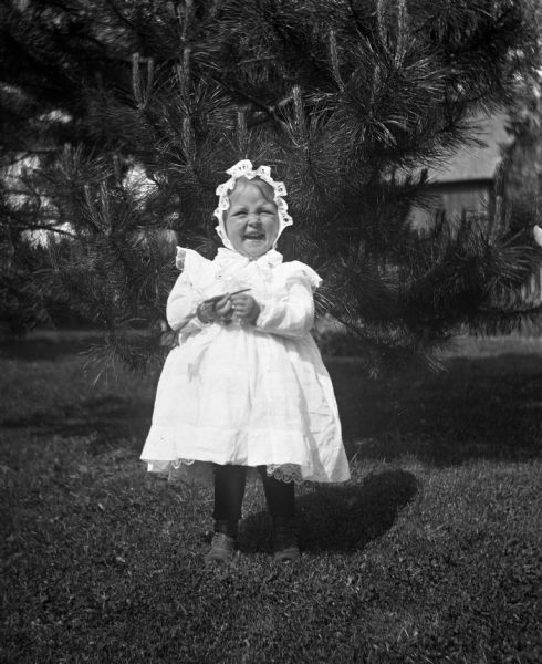 Outdoor portrait of Mable Klosowsky standing in front of a small pine tree. She is holding a flower in her hand while laughing. In the background is a barn among trees.
