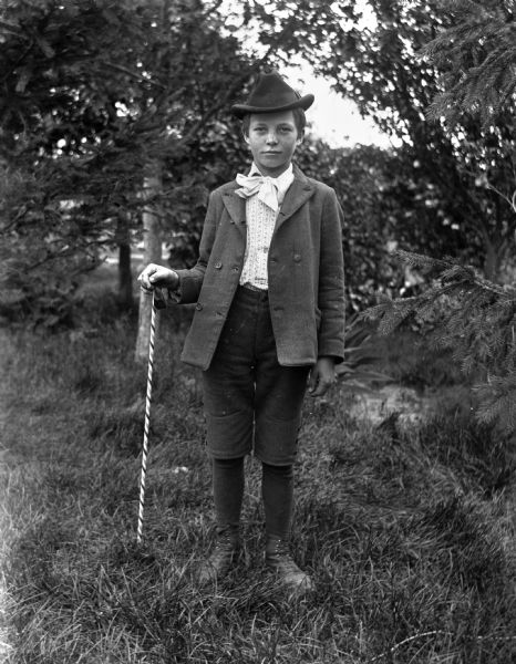 Outdoor portrait of an unidentified boy standing in the grass among pine trees. He is holding a striped walking stick in his right hand.