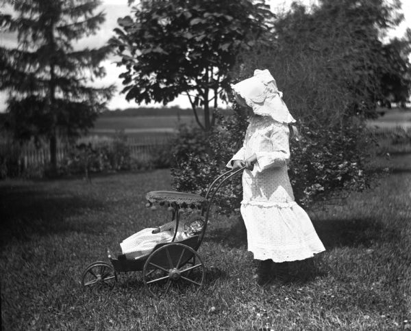 An unidentified girl wearing a bonnet pushing her doll in a small stroller across the lawn.