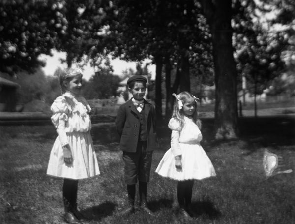 Outdoor portrait of three unidentified children standing in a yard. The two young girls are wearing white dresses with puffy sleeves. The young boy is wearing a suit and hat. Buildings are in the far background.