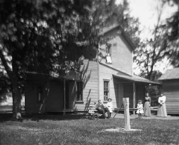 View across yard of an unidentified family posing in front of their home. In the foreground is a hand-pump. From the left a young boy is sitting in a rocking chair, a man is sitting in a large wood chair, and two young girls and two women are standing in the center on the right. Behind them is the back porch of a house and an outbuilding.