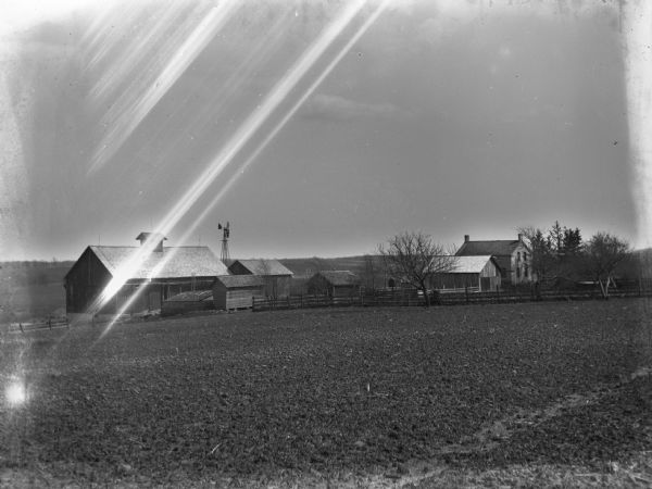 View across field of a farmhouse, a windmill, and farm buildings.