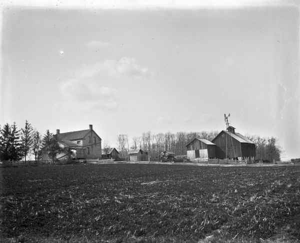 View across cleared field of a farmhouse, windmill and farm buildings.
