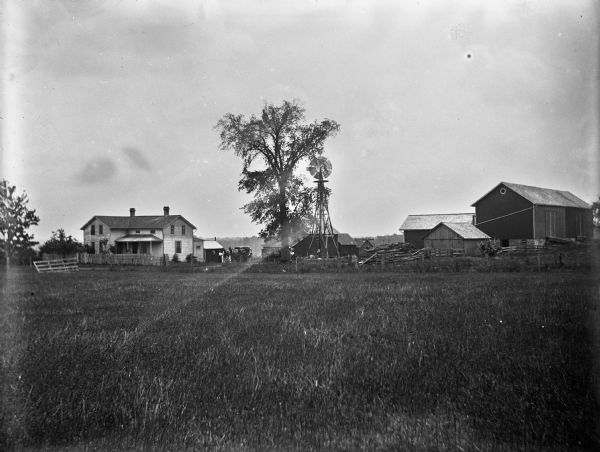 View across field towards a farmhouse, windmill and farm buildings. Several people are standing near a number of horse-drawn carriages near the farmhouse.