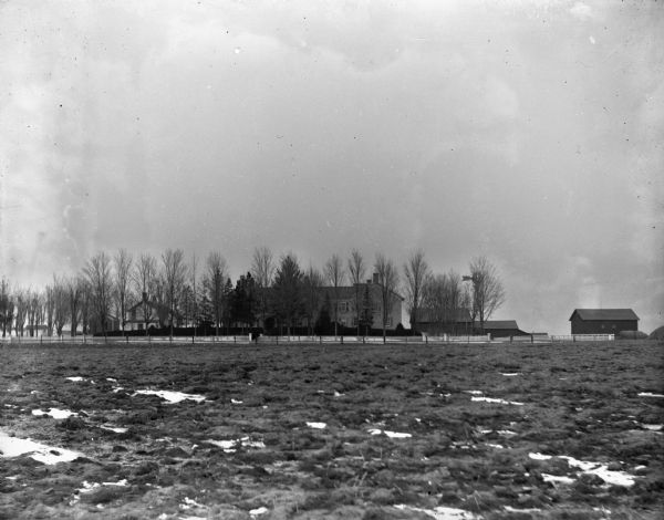 View across field with patches of snow towards a farmstead behind a row of trees.