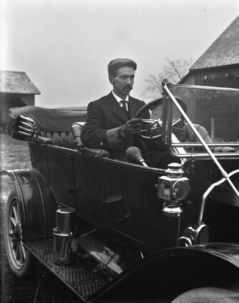 August Kressin sitting in the driver's seat of a Flanders 20. He is wearing a pair of driving gloves. Farm buildings are in the background.
