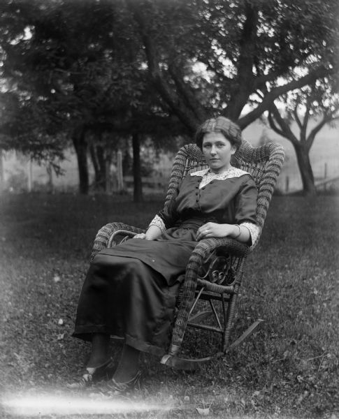 Outdoor portrait of Jennie Krueger Bruetzman sitting in a wicker rocking chair on the Krueger farm. Behind her are trees, a fence, and fields.