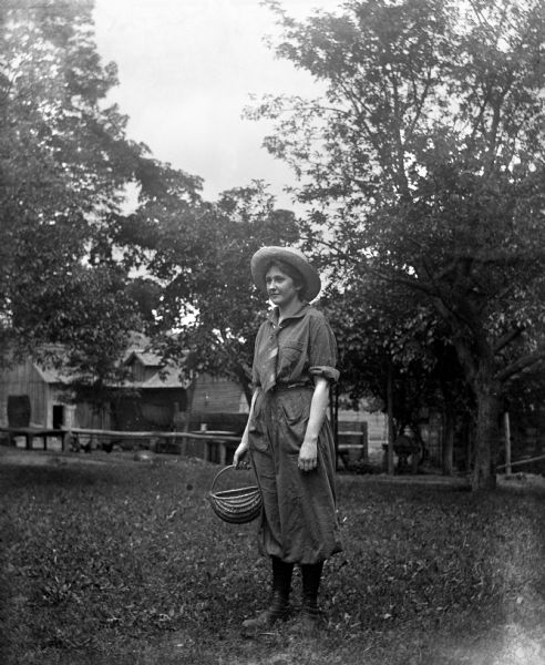 Jennie Krueger Bruetzman standing in the yard on the Krueger farm. She is wearing pants and holding a basket. Chickens are near farm buildings in the background.