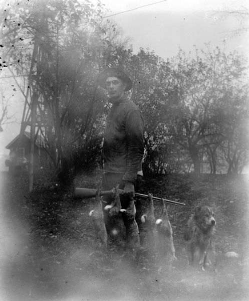 Edgar Krueger posing with five rabbits he shot, which are hanging by their feet from the rifle in his hand. A dog is sitting next to him. In the background on the left is a tall structure, possibly a windmill, and a farm building.
