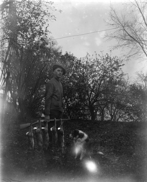Edgar Krueger posing with five rabbits he shot. The rabbits are hanging by their feet from the rifle he is holding down at his side. A dog is sitting next to the rabbits. In the background on the left is a tall structure, possibly a windmill, and a farm building.