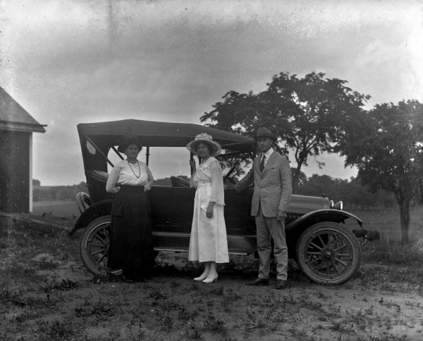 Florentina, Jennie, and Edgar Krueger posing next to a 1917 Chevrolet touring car on the Krueger farm. In the background, cows or horses are grazing in a field. There is part of a building on the left.