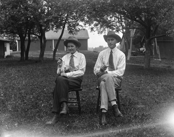 Outdoor portrait of Jennie and Edgar Krueger sitting in chairs on the Krueger farm. Both of them are wearing hats, neckties, suit's, and are smoking cigars.