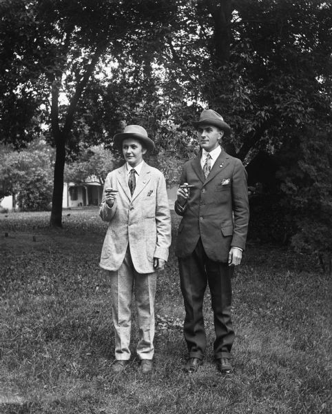 Outdoor portrait of Jennie and Edgar Krueger. They are both smoking cigars and wearing suits, hats, and ties.