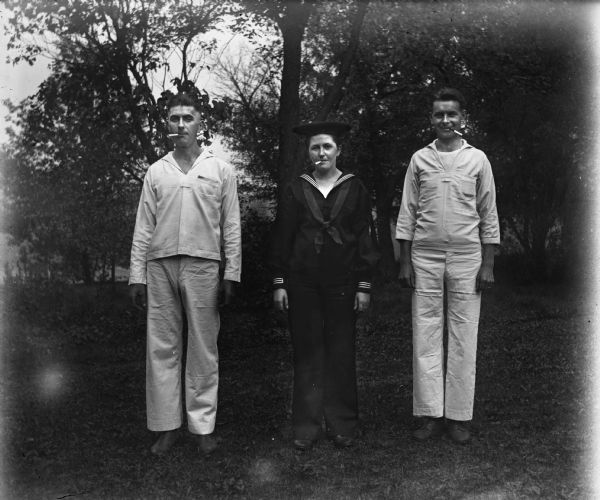 Outdoor portrait of Edgar Krueger, Jennie Krueger Bruetzman, and Ernst Bruetzman in sailor uniforms with cigarettes in their mouths. Edgar and Ernst are in white uniforms while Jennie is in a blue US Navy uniform.