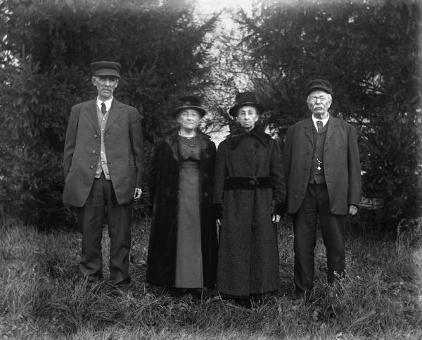 Outdoor group portrait of August Buelke, Martha Sara Goetsch Buelke, Mary Goetsch Krueger, and August Krueger standing by a line of pine trees.