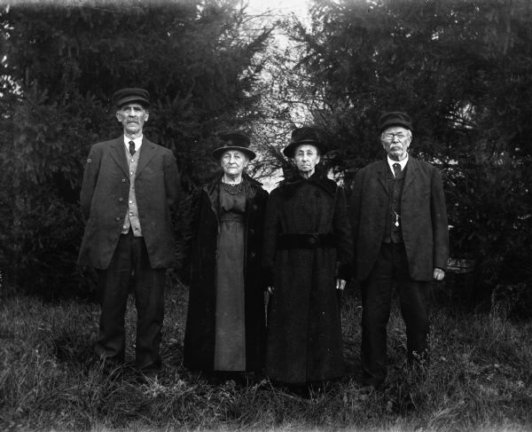 Outdoor group portrait of August Buelke, Martha Buelke, Mary Krueger, and August Krueger posing together in front of a row of pine trees.