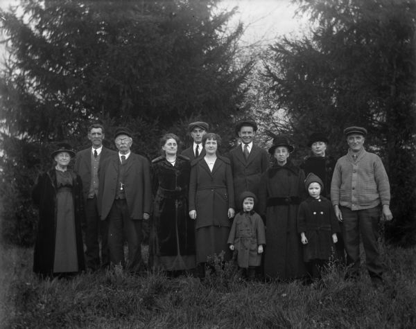 Outdoor group portrait of the Kruegers, Bhends, and Buelkes in front of a row of pine trees. From left to right: Martha Goetsch Buelke, August Buelke, August Krueger, Florentina Krueger, Edgar Krueger, Jennie Krueger Bruetzman, Ernst Bruetzman, Mary Krueger, Sara Krueger Bhend, John Bhend, and the two Bhend children (Irene and Marcel).