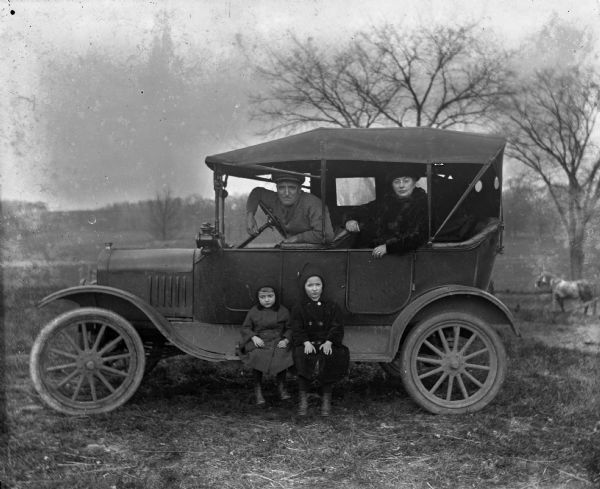 The Bhend family posing with their first car, a Ford Model T, on the Krueger farm. Posing and looking out of the car windows are John Bhend, sitting in the driver's seat, and Sara Krueger Bhend sitting in the backseat. Their two children, Irene and Marcel, are sitting below them on the running board. In the background is a horse in a field.