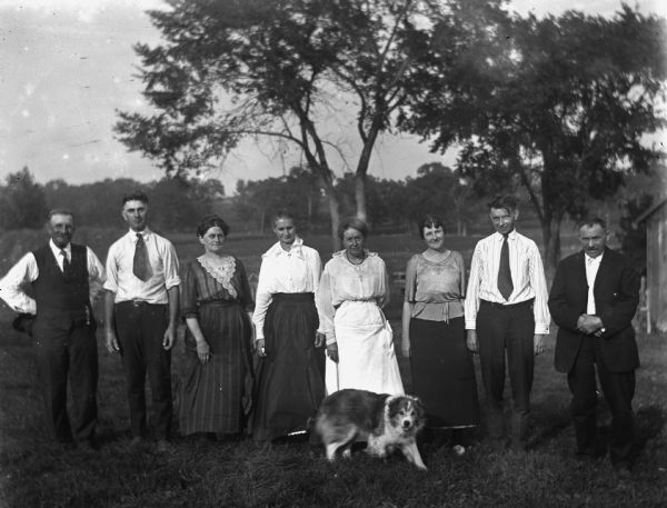 Outdoor group portrait of the Kruegers, Zemkes, and Bruetzmans standing in a line, with a dog standing in front of them. From left to right William Zemke, Edgar Krueger, Florentina Krueger, Mrs. August Bruetzman, Emma Zemke, Jennie Krueger Bruetzman, Ernst Bruetzman, and August Bruetzman.