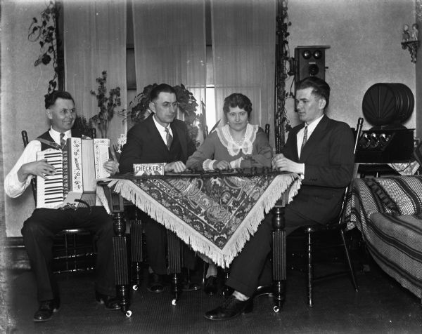 Indoor flashlight portrait of Ernst Bruetzman, Edgar Krueger, Jennie Krueger Bruetzman, and Raymond Fehrmann sitting at the table. Ernst is playing the accordion while the others are playing checkers. A telephone and large radio are in the background.