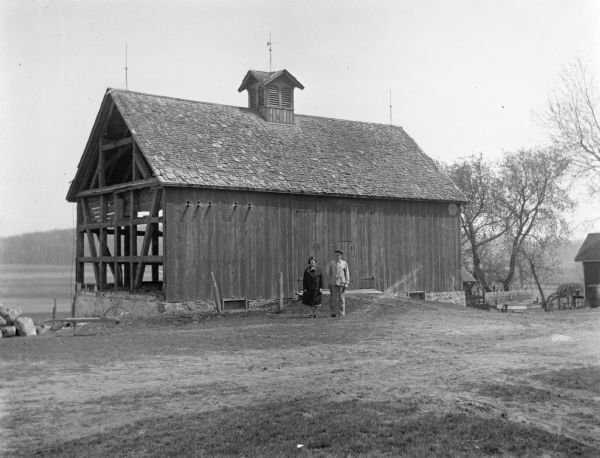 Alexander and Florentina Krueger posing together in front of an old barn on Krueger farm. The barn was being torn down and parts of the framework are showing.