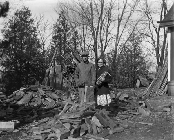 Edgar and Elna Krueger posing together next to a pile of firewood. Edna is holding one of the pieces in her hand while Edgar is holding an axe and another piece of firewood.