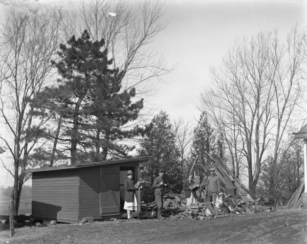 Florentina, Alexander, Elna, and Edgar Krueger are standing next to a wood pile with two of their dogs. A small wagon shed is to the left of the pile. Florentina and Elna are each holding a piece of the cut wood, while Edgar is holding an axe that rests on an upright wood piece.