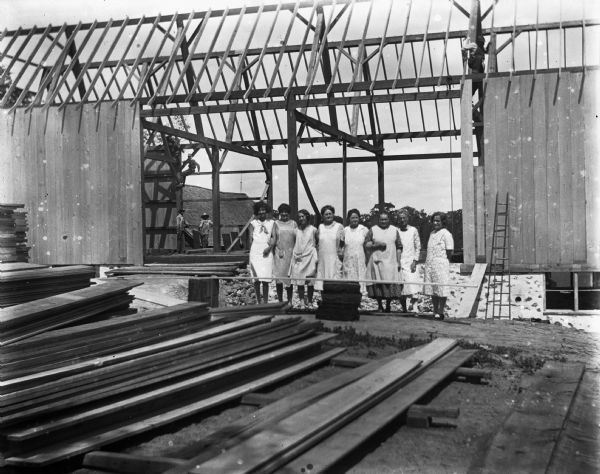 Group of women posing in front of the new barn foundation. Jennie Krueger is standing second from the right, Florentina Krueger is standing fourth from the right, and Elna Krueger is standing on the far left. Several men are working on the barn in the background. The women came together to make food for the working men.