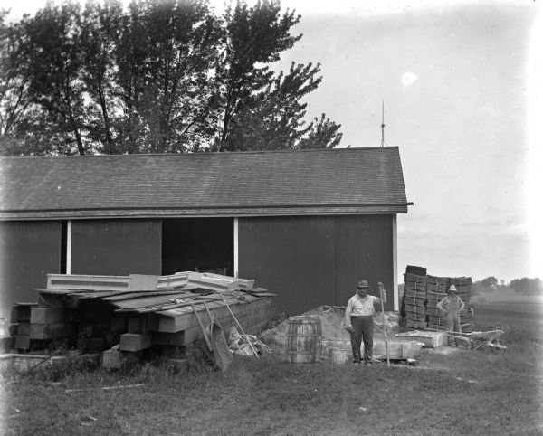 Two men mixing mortar for the building of the barn. They are standing next to a large pile of wood in front of a shed.