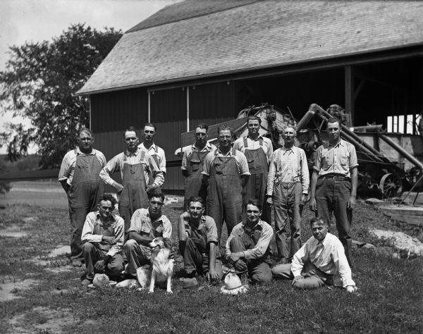 A group of thirteen men are standing in front of a newly constructed barn. Alexander Krueger is standing in the back, second from left, and Edgar Krueger is standing in back third from the right. A dog is sitting in front of the men.